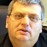 גבר בגיל העמידה עם שיער אפור קצר ומשקפיים מביט לעבר המצלמה, יושב בתוך חדר עם ריהוט משרדי ברקע.