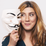 אישה עם שיער ארוך וחום בהיר מחזיקה מסכת גיא פוקס ליד פניה, מביטה בה בחיוך קל על רקע אחיד.