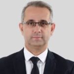 גבר עם שיער אפור קצר, המרכיב משקפיים, לבוש בחליפה כהה, חולצה לבנה ועניבה שחורה, מצטלם על רקע אפור בהיר.